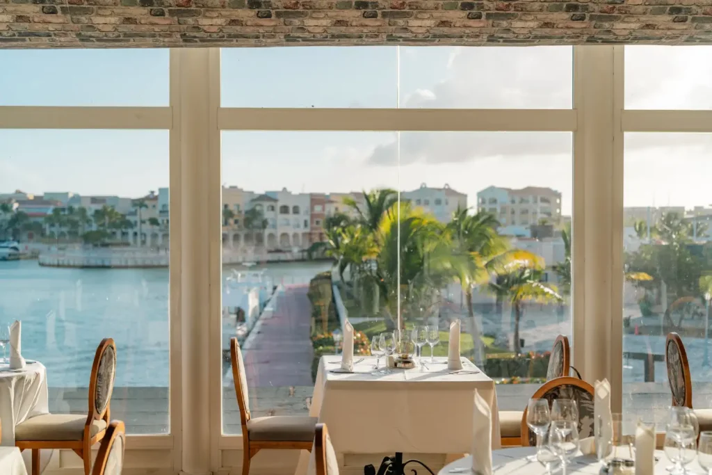 Restaurant dining room with waterfront view, palm trees, and formal table setup.