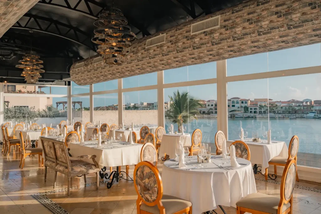 Dining room with round tables, white linens, and waterfront views through large windows.