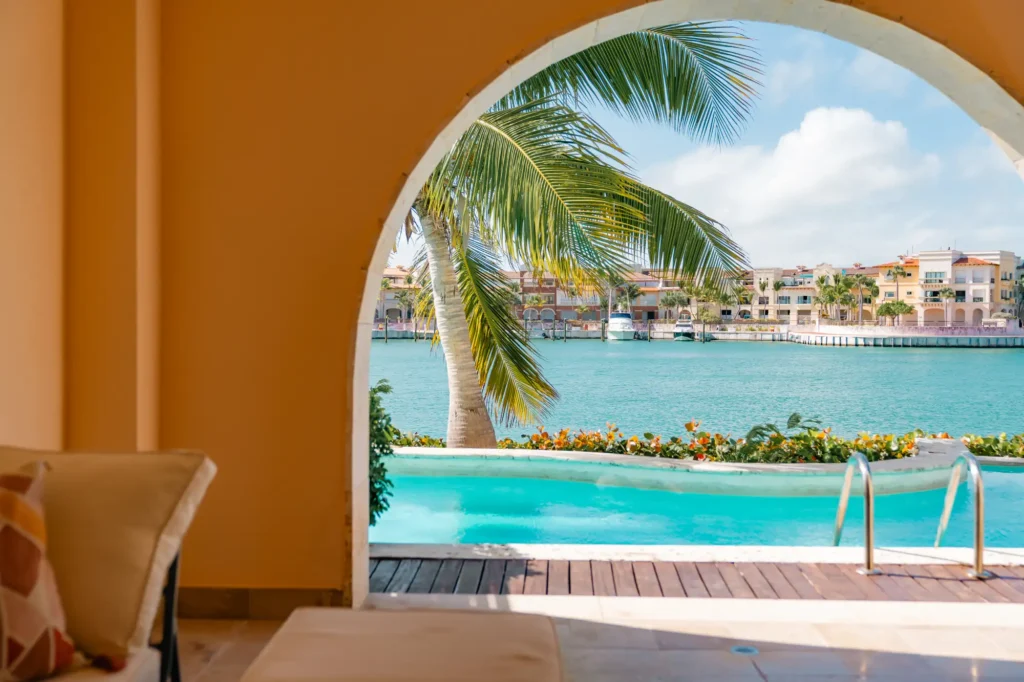 Pool and turquoise water view through arched opening with palm tree fronds visible.