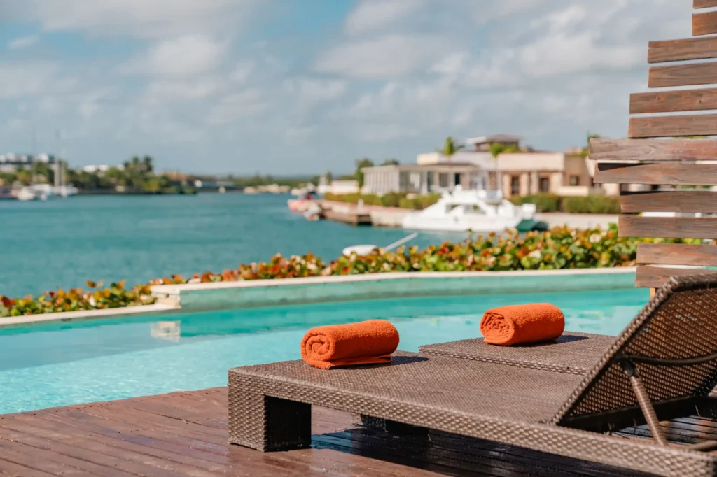 Pool deck with lounger, rolled towels, waterfront view, and tropical landscaping.