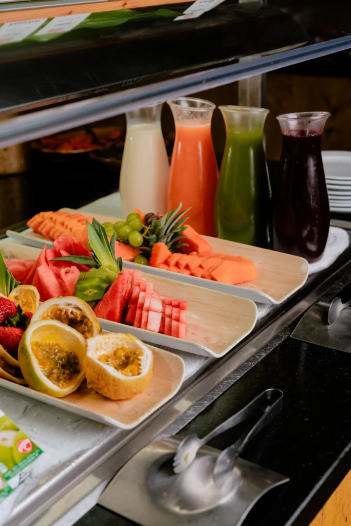 A buffet station with several trays of fresh-cut tropical fruits like passion fruit, watermelon, and papaya, alongside four glass carafes of different colored juices.