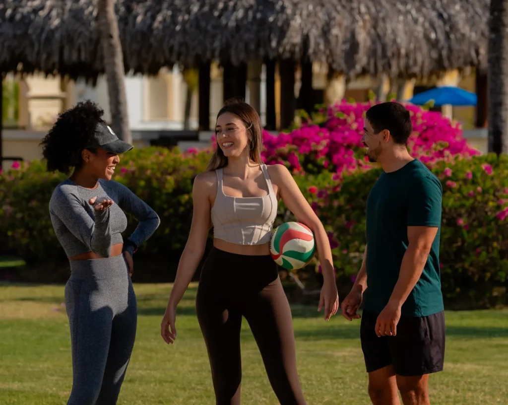 Three people in athletic wear stand on a grassy lawn at a tropical resort, talking and smiling while one woman holds a volleyball.