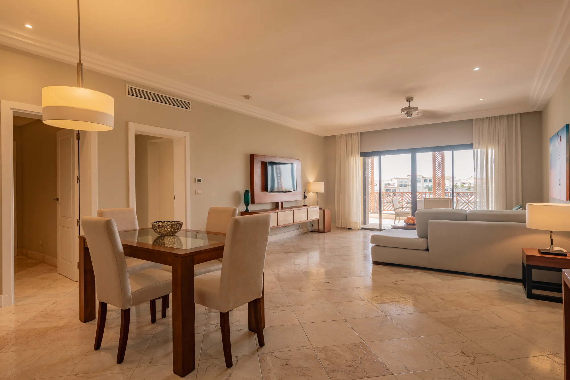 A spacious, open-plan hotel suite featuring a dining area with a four-seat table and a living area with a large sectional sofa, tiled floors, and a balcony.